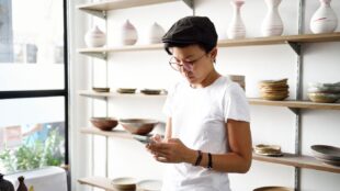 Propriétaire d'une petite entreprise regardant son téléphone dans un studio avec des étagères remplies de poteries artisanales derrière lui.
