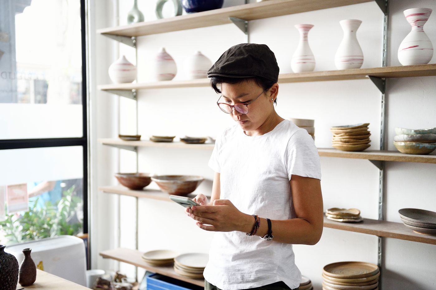 Small business owner looking at their phone in a studio with shelves of handmade pottery behind them.