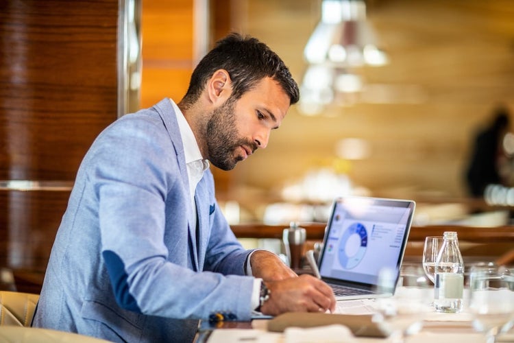 Man writing and reviewing pie chart on laptop