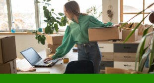 Woman holding cardboard box while operating her computer with other hand.
