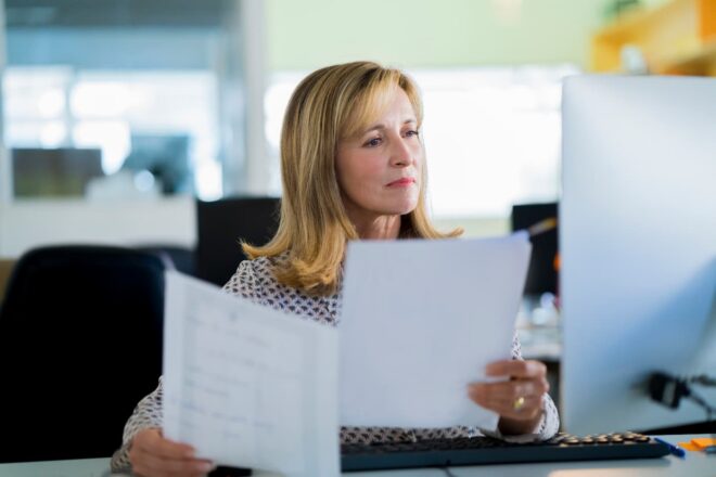 Professional woman holding papers and looking at monitor