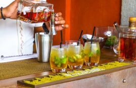 Happy hour menu drinks placed in a row on a bar top while a bartender prepares another cocktail by mixing ingredients.