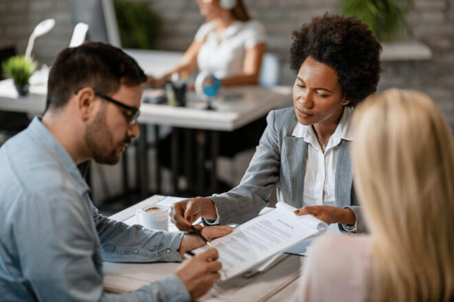 Female real estate agent sitting with a couple in an office while reading over a lease agreement with the couple.