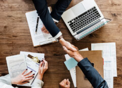 Two men's arms reaching across a table, shaking hands making a business deal. Woman's hands are also seen resting on a pile of business papers with a pen in her hand next to one of the man's arms.