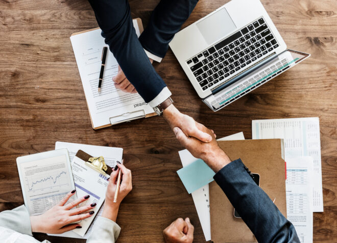 Two men's arms reaching across a table, shaking hands making a business deal. Woman's hands are also seen resting on a pile of business papers with a pen in her hand next to one of the man's arms.