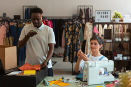 Man standing in a clothing store taking a picture of a shirt while woman sits next to man using a sewing machine.