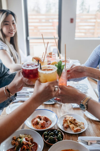 Group of friends sitting at a table holding happy hour drinks in the center of the table.